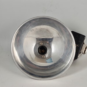 May include: A vintage photographic flash unit with a large, reflective silver bowl-shaped reflector. The flash head has a black control panel with dials and a connector. The reflector is made of polished metal.