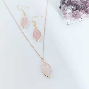 May include: A rose gold jewelry set featuring a necklace and earrings. The necklace has a delicate chain with a teardrop-shaped pendant. The earrings are matching teardrop-shaped dangles. Each piece has a pale pink gemstone set in a gold frame.