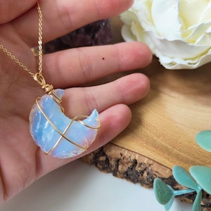 May include: A gold-coloured wire-wrapped necklace featuring a crescent moon-shaped opalite stone. The opalite has a translucent, milky white appearance with hints of pink and blue. The necklace is displayed against a wooden surface.