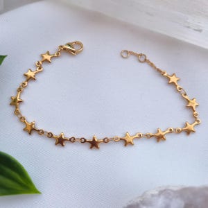 May include: Gold-coloured bracelet with star-shaped charms. The bracelet has a delicate chain and a clasp closure. The stars are evenly spaced along the chain, creating a celestial theme. The bracelet is displayed on a white surface.