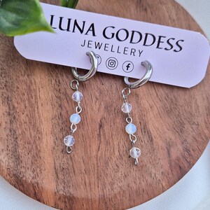 K&ouml;nnte beinhalten: Silberne Creolen-Ohrringe mit herabh&auml;ngenden, schillernden Perlen. Die Ohrringe werden auf einer Holzoberfl&auml;che mit einem Etikett "LUNA GODDESS JEWELLERY" pr&auml;sentiert. Das Design ist zart und elegant.