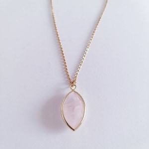 May include: A gold-colored chain necklace with a pendant. The pendant is a light pink, oval-shaped stone set in a gold-colored frame. The necklace is displayed against a white background.