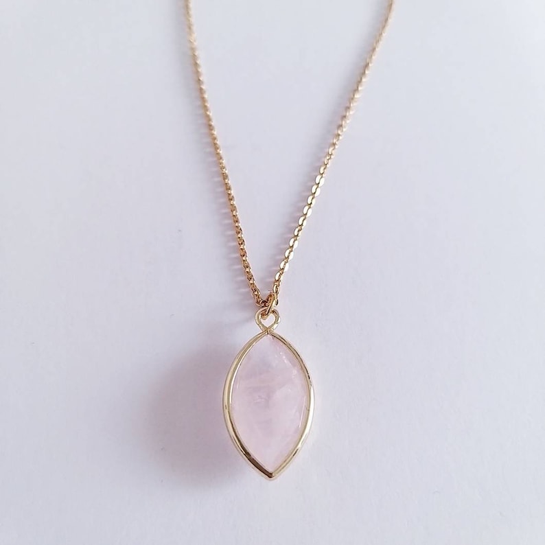 May include: A gold-colored chain necklace with a pendant. The pendant is a light pink, oval-shaped stone set in a gold-colored frame. The necklace is displayed against a white background.