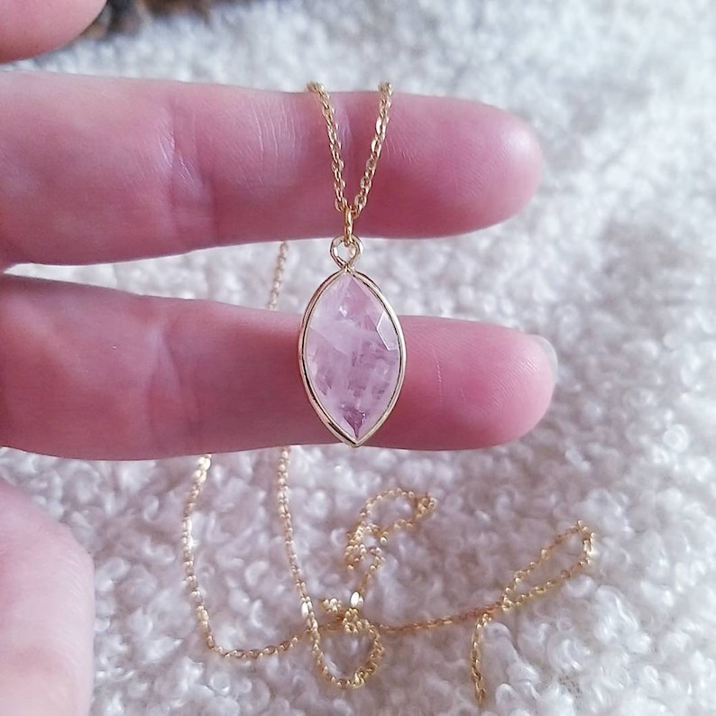 May include: A gold-colored necklace with a pendant featuring a faceted, light pink gemstone. The pendant is an elongated oval shape, set in a gold-colored bezel. The chain is delicate and also gold-colored, creating an elegant jewelry piece.