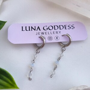 K&ouml;nnte beinhalten: Silberne Creolen-Ohrringe mit herabh&auml;ngenden Str&auml;ngen aus kleinen, klaren und opalisierenden Perlen. Die Ohrringe werden auf einem hellvioletten Etikett mit der Aufschrift "LUNA GODDESS JEWELLERY" pr&auml;sentiert. Der Hintergrund ist ein weicher, wei&szlig;er Stoff.