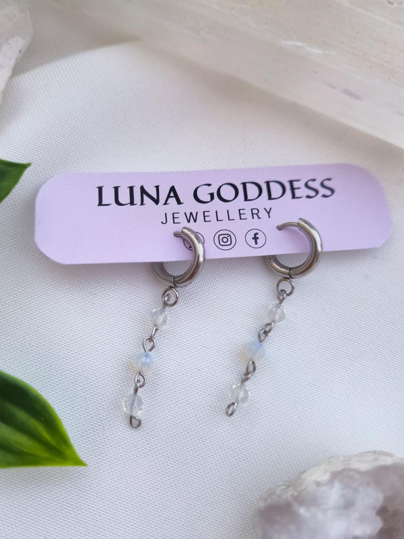 K&ouml;nnte beinhalten: Silberne Creolen-Ohrringe mit herabh&auml;ngenden Str&auml;ngen aus kleinen, klaren und opalisierenden Perlen. Die Ohrringe werden auf einem hellvioletten Etikett mit der Aufschrift "LUNA GODDESS JEWELLERY" pr&auml;sentiert. Der Hintergrund ist ein weicher, wei&szlig;er Stoff.