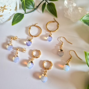 May include: A collection of gold-colored earrings with iridescent, teardrop-shaped pendants. The earrings include hoop styles and stud earrings. The pendants have a pearlescent, opalescent appearance. The earrings are displayed on a white surface.
