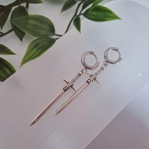 May include: A pair of silver earrings with a sword design. Each earring has a hoop closure and a detailed sword pendant. The earrings are displayed on a white surface with green foliage in the background.