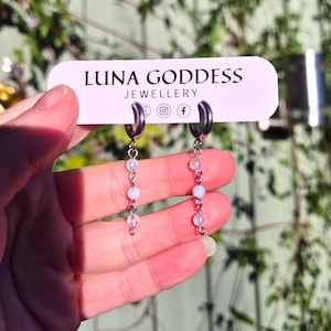 K&ouml;nnte beinhalten: Silberne Creolen-Ohrringe mit herabh&auml;ngenden Str&auml;ngen aus kleinen, schillernden Perlen. Die Ohrringe werden vor einem Schild mit der Aufschrift "LUNA GODDESS JEWELLERY" pr&auml;sentiert. Der Hintergrund ist verschwommen mit gr&uuml;nem Laub.