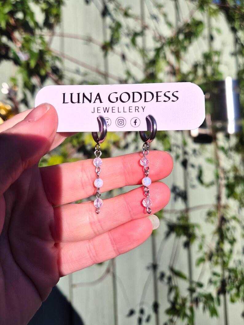 K&ouml;nnte beinhalten: Silberne Creolen-Ohrringe mit herabh&auml;ngenden Str&auml;ngen aus kleinen, schillernden Perlen. Die Ohrringe werden vor einem Schild mit der Aufschrift "LUNA GODDESS JEWELLERY" pr&auml;sentiert. Der Hintergrund ist verschwommen mit gr&uuml;nem Laub.