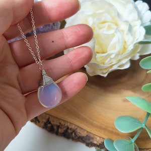May include: A silver necklace with a teardrop-shaped pendant. The pendant is a pale, opalescent stone wrapped in silver wire. The chain is delicate and silver. The background includes a white flower and greenery.