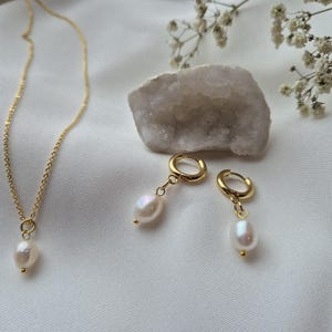 Dainty pearl necklace on 18k gold plated chain with matching higgie hoop earrings on a soft white fabric background surrounded by flowers and crystals