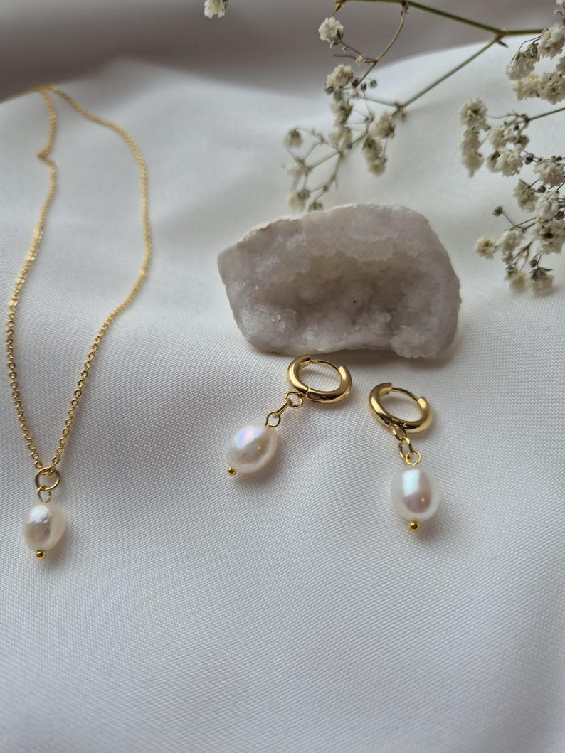 Dainty pearl necklace on 18k gold plated chain with matching higgie hoop earrings on a soft white fabric background surrounded by flowers and crystals