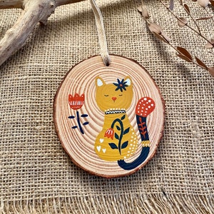 Peut inclure: Ornement rond en bois avec un motif de chat peint. Le chat est jaune avec une fleur bleue et une queue tachetée rouge et blanche. Une fleur rouge et des feuilles bleues sont peintes à gauche. Une boucle en ficelle est attachée pour accrocher.