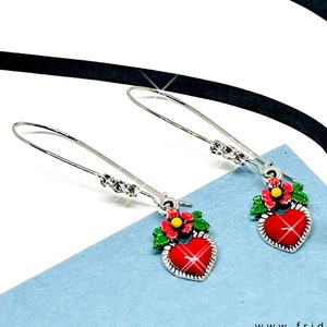Peut inclure: Une paire de boucles d'oreilles pendantes argent&eacute;es avec des breloques en forme de c&oelig;ur rouge. Chaque breloque en forme de c&oelig;ur est surmont&eacute;e d'une feuille verte et d'une fleur rouge et jaune. Les boucles d'oreilles sont sur une surface bleue avec un ruban noir et des fleurs s&eacute;ch&eacute;es en arri&egrave;re-plan.