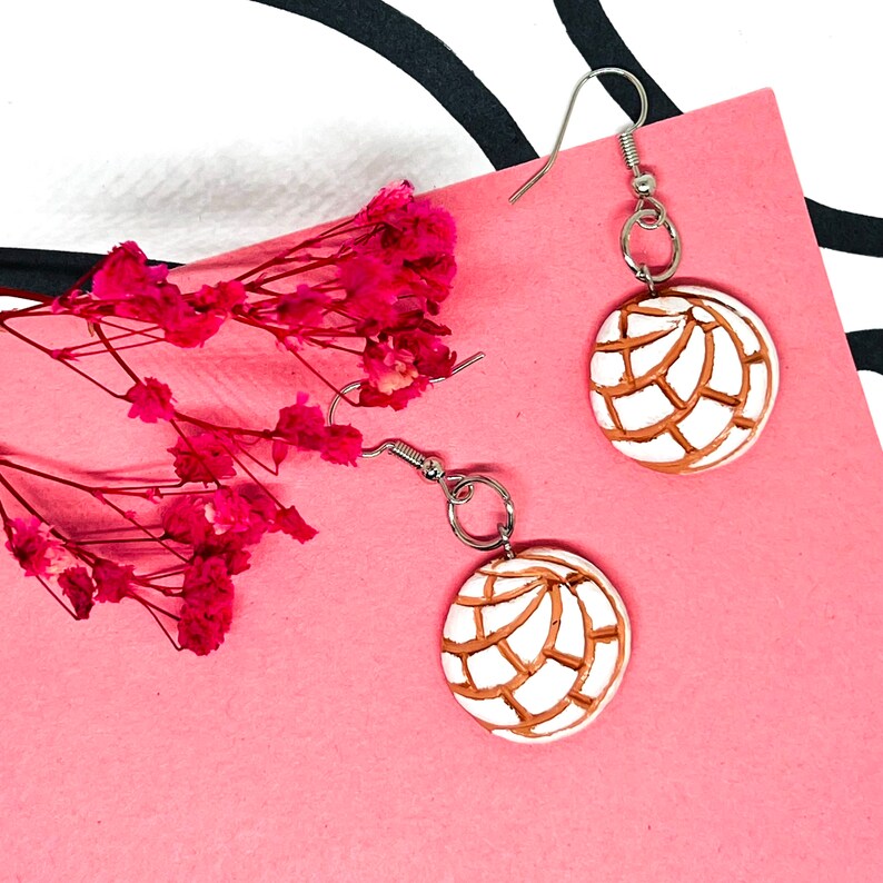 Puede incluir: Un par de aretes colgantes de plata con un dije redondo blanco y marr&oacute;n. El dije es una r&eacute;plica de un pan dulce mexicano llamado concha.