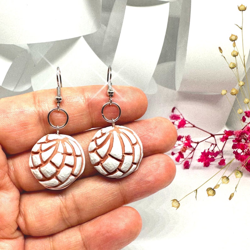 Puede incluir: Un par de aretes colgantes de plata con dijes de arcilla blanca y marr&oacute;n con forma de concha, un pan dulce mexicano tradicional.