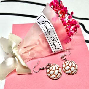 Puede incluir: Un par de aretes colgantes con un dise&ntilde;o en forma de concha blanco y marr&oacute;n. Los aretes cuelgan de ganchos plateados. Los aretes se muestran sobre una superficie rosa con un ramo de flores rosas y una etiqueta blanca que dice "Eternal Angel".