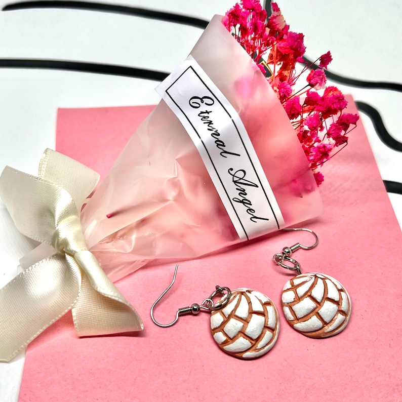 Puede incluir: Un par de aretes colgantes con un dise&ntilde;o en forma de concha blanco y marr&oacute;n. Los aretes cuelgan de ganchos plateados. Los aretes se muestran sobre una superficie rosa con un ramo de flores rosas y una etiqueta blanca que dice "Eternal Angel".