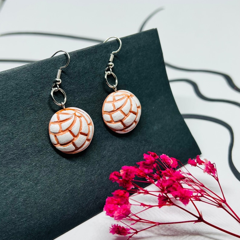 Puede incluir: Un par de aretes colgantes de plata con dijes de arcilla blanca y naranja con forma de pan dulce mexicano, concha.