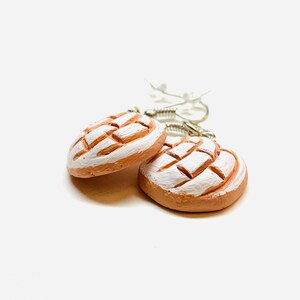 Puede incluir: Un par de pendientes colgantes con un dise&ntilde;o de cer&aacute;mica esmaltada blanca y marr&oacute;n que se asemeja a un pan dulce.