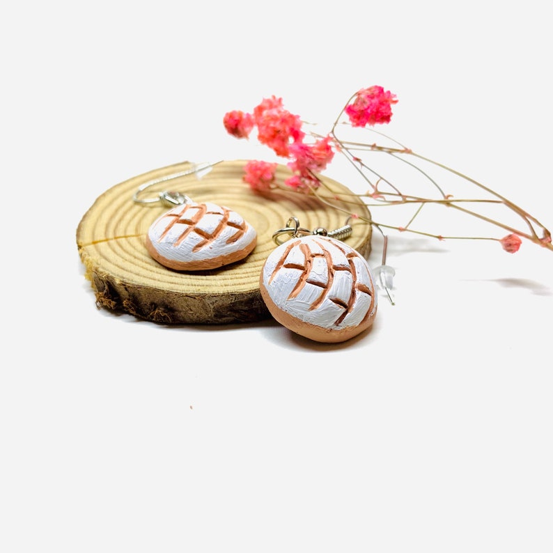 Puede incluir: Un par de pendientes colgantes con un dise&ntilde;o de pan concha blanco y marr&oacute;n. Los pendientes se muestran sobre una superficie de madera con flores rosas secas.