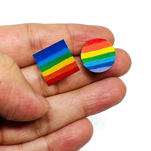 May include: Two wooden rainbow-colored earrings, one square and one round, are shown in a hand.