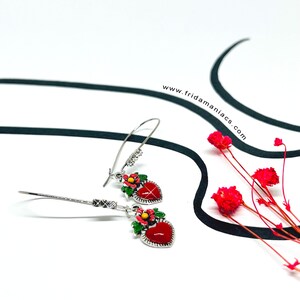 Peut inclure: Une paire de boucles d'oreilles pendantes argent&eacute;es avec des pendentifs en forme de c&oelig;ur rouge. Chaque pendentif en forme de c&oelig;ur a un motif floral vert et une finition &eacute;maill&eacute;e rouge. Les boucles d'oreilles sont sur un fond blanc.