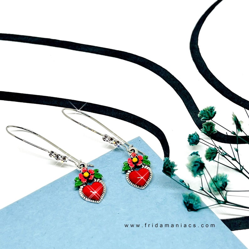 Peut inclure: Une paire de boucles d'oreilles pendantes argent&eacute;es avec des breloques en forme de c&oelig;ur rouge. Chaque breloque en forme de c&oelig;ur est surmont&eacute;e d'une feuille verte et d'une fleur rouge et jaune. Les boucles d'oreilles sont sur une surface bleue avec un ruban noir et des fleurs s&eacute;ch&eacute;es en arri&egrave;re-plan.
