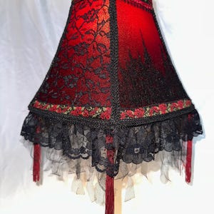 Peut inclure: Abat-jour rouge et noir avec un dessus carré et des côtés évasés. L'abat-jour est recouvert de dentelle noire sur du tissu rouge. Le bord inférieur présente une garniture en dentelle noire avec des glands rouges. Le bord supérieur a une garniture florale.