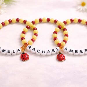 Könnte beinhalten: Drei Armbänder mit den Namen "PAMELA", "RACHAEL" und "AMBER". Jedes Armband hat gelbe und rote Perlen, weiße Buchstabenperlen und einen Marienkäfer-Anhänger. Sie liegen auf einer weißen Oberfläche.
