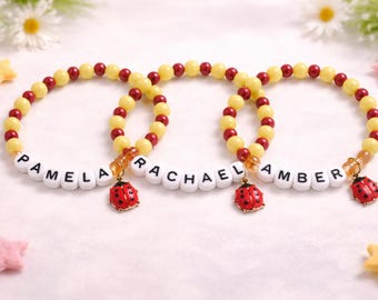 Könnte beinhalten: Drei Armbänder mit den Namen "PAMELA", "RACHAEL" und "AMBER". Jedes Armband hat gelbe und rote Perlen, weiße Buchstabenperlen und einen Marienkäfer-Anhänger. Sie liegen auf einer weißen Oberfläche.