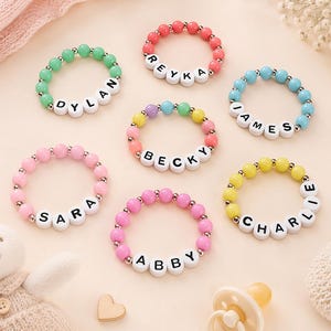 May include: A selection of colourful beaded bracelets, each spelling out a name with white letter beads. The bracelets are in shades of green, pink, red, blue, yellow, and mixed colours. Names include Dylan, Reyka, James, Becky, Sara, Abby, and Charlie.