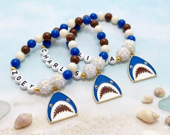 May include: Three beaded bracelets with blue, white, and brown beads. Each bracelet has a shark charm and a name spelled out in white letter beads. The shark charms are blue with a white border and a detailed mouth.