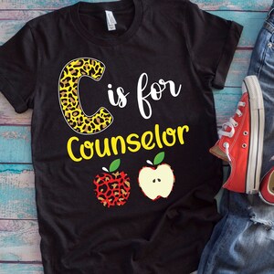 May include: Black t-shirt with the text "C is for Counselor" in white and yellow. The letter "C" has a leopard print design. Two apple graphics are below the text. The shirt is paired with blue jeans and red sneakers.