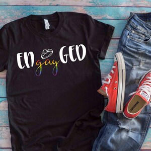 Könnte beinhalten: Schwarzes T-Shirt mit dem Text "Engaged" und einer Ringgrafik. Das Wort "gay" ist in Regenbogenfarben gehalten. Das Shirt wird mit blauen Jeans und roten Converse-Sneakern kombiniert.
