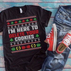 Puede incluir: Camiseta negra con un diseño de suéter navideño blanco con el texto "Estoy aquí para eliminar tus cookies". La camiseta se combina con jeans azules y zapatillas Converse rojas.
