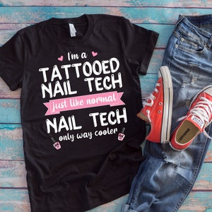 Könnte beinhalten: Schwarzes T-Shirt mit pinkfarbenem Text, der "I'm a Tattooed Nail Tech Just like normal Nail Tech only way cooler" lautet. Das Shirt liegt auf einer blauen Holzoberfläche mit einer blauen Jeans und roten Converse-Schuhen.