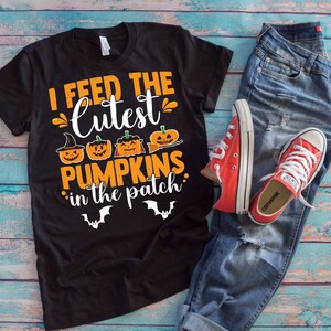 May include: Black t-shirt with the text "I feed the cutest pumpkins in the patch" in orange and white. The shirt features pumpkin graphics and bat silhouettes. The shirt is paired with blue jeans and red Converse sneakers.