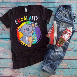 Könnte beinhalten: Schwarzes T-Shirt mit einem Regenbogen und einem Cartoon-Koala, der an einem Baumzweig festhält. Der Text "EKOALATTY" ist auf das Shirt gedruckt.
