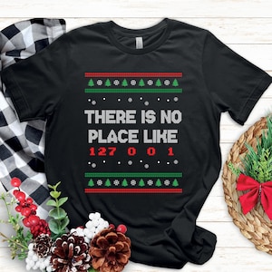 Könnte beinhalten: Schwarzes T-Shirt mit festlichem Design. Das Shirt zeigt den Text "THERE IS NO PLACE LIKE 127 001" in Weiß und Rot, mit grünen und roten Weihnachtsbaum- und Schneeflockenmustern oben und unten. Ein Shirt zum Thema Feiertage.