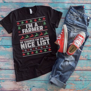 Pode incluir: T-shirt preto com o texto "I'M A FARMER OF COURSE I'M ON THE NICE LIST" em branco, vermelho e verde. A camisa é combinada com jeans azuis e tênis vermelhos Converse. O design inclui árvores de Natal e renas.