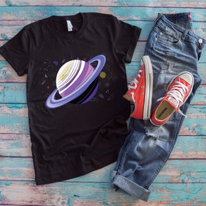 Peut inclure: Un t-shirt noir avec un motif de planète violette et blanche avec des anneaux dans une scène spatiale. Le t-shirt est posé sur une surface en bois bleu et blanc avec une paire de chaussures Converse rouges et un jean bleu.