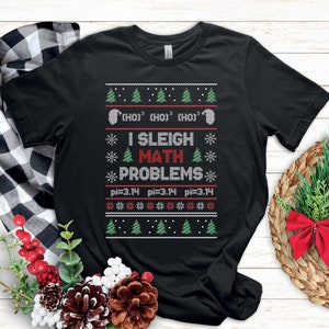 May include: Black t-shirt with a festive design. The shirt features the text "I Sleigh Math Problems" in red and green, along with mathematical symbols and Christmas tree graphics. The shirt also includes the pi symbol and the number 3.14.