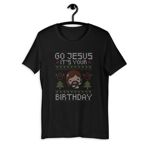May include: Black t-shirt with a festive design. The shirt features the text "GO JESUS IT'S YOUR BIRTHDAY" in a pixelated font, along with images of a cartoon Jesus, snowmen, and Christmas trees. The design has red and green accents.