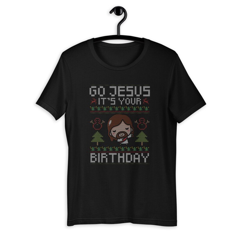 May include: Black t-shirt with a festive design. The shirt features the text "GO JESUS IT'S YOUR BIRTHDAY" in a pixelated font, along with images of a cartoon Jesus, snowmen, and Christmas trees. The design has red and green accents.