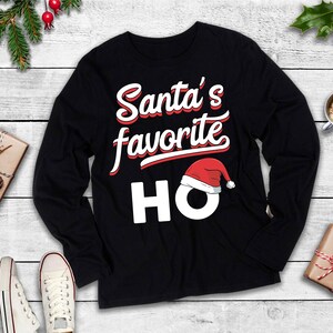 May include: Black long-sleeved top featuring the text "Santa's favourite HO" in white and red. A Santa hat graphic sits above the letters "HO". The shirt is presented on a white wooden surface with festive decorations.
