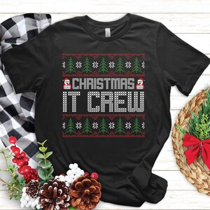 May include: Black t-shirt with a festive design. The shirt features the text "CHRISTMAS IT CREW" in white, surrounded by a pattern of green Christmas trees, red snowflakes, and Santa Claus figures. A holiday-themed apparel item.