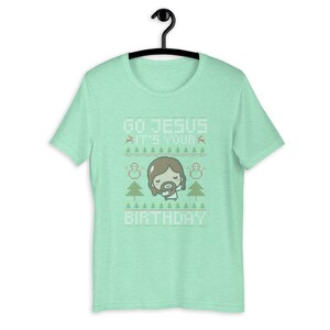 May include: A light green t-shirt with the text "GO JESUS IT'S YOUR BIRTHDAY" in white. The design includes a cartoon Jesus, snowmen, Christmas trees, and reindeer. The shirt is hanging on a black hanger.
