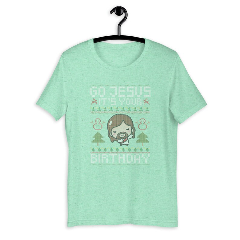 May include: A light green t-shirt with the text "GO JESUS IT'S YOUR BIRTHDAY" in white. The design includes a cartoon Jesus, snowmen, Christmas trees, and reindeer. The shirt is hanging on a black hanger.